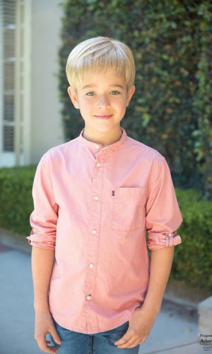 Young boy with blonde hair in a pink shirt outdoors, smiling at the camera.