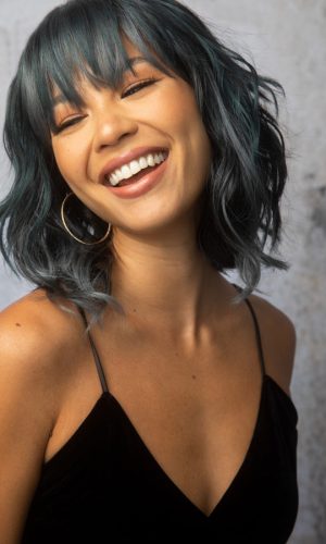 Woman with wavy black hair and bangs, smiling and wearing hoop earrings.