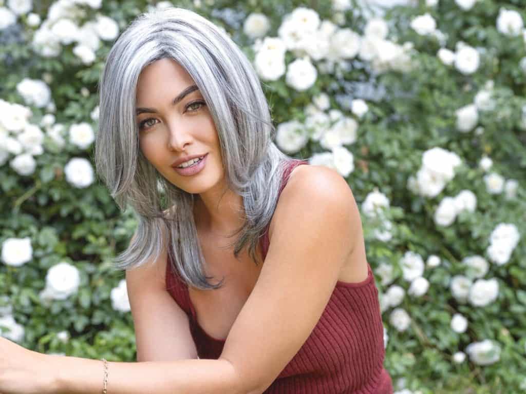 Woman with gray wavy wig outdoors among white flowers.