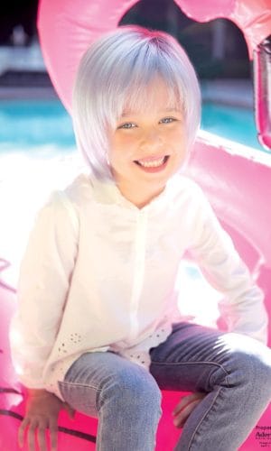 Child with silver wig smiling on pink float in pool.