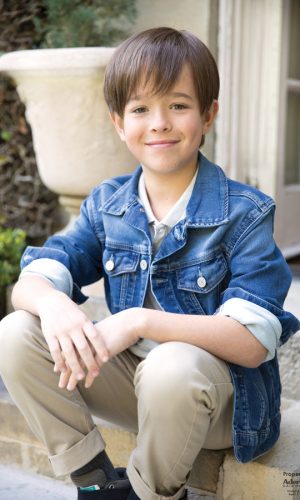Young boy wearing a short brown wig, denim jacket, and beige pants outdoors.