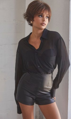 Model with short curly hair wearing black blouse and leather shorts.