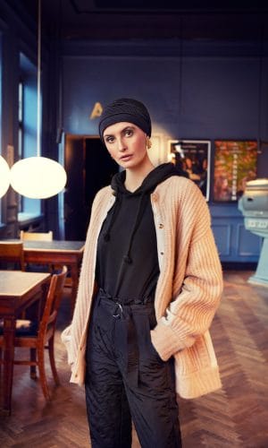 Young woman wearing a braided turban wig, black hoodie, and beige cardigan in a cafe.