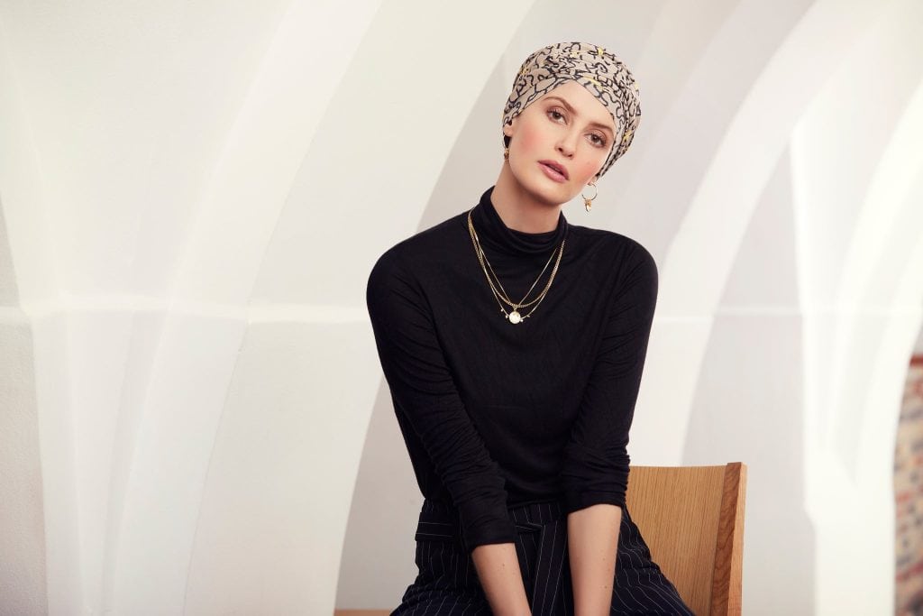 Woman wearing a patterned turban and black top, sitting on a wooden chair.