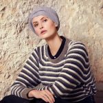 Woman wearing a gray turban and striped sweater against a textured wall.