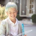Child wearing a pastel-colored wig smiling outdoors in sunlight.