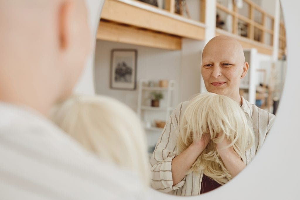 Woman with bald head examining a blonde wig in a mirror at Hidden Secrets Wigs.