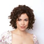Woman with curly wig, natural makeup, floral top, looking at camera.