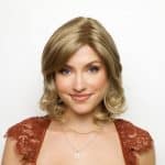 Blonde wig styled on a woman with soft waves, natural makeup, and a rust-colored dress.