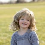 Adorable young girl with wavy blonde hair smiling outdoors in a grassy field.