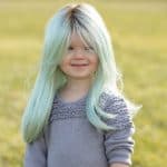 Girl with mint green wig outdoors, smiling, in a grassy field.