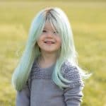 Young girl wearing a light blue wig outdoors, smiling happily in a grassy field.