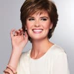 Woman with short layered brown wig smiling and touching her ear.
