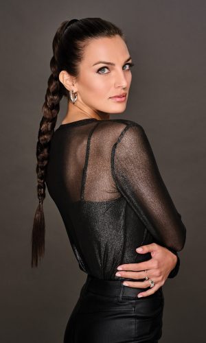 Woman with long braided wig in a black sheer top and dark background.
