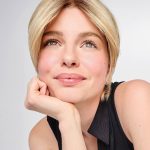 Blonde woman with short layered wig, smiling gently, resting her chin on her hand.