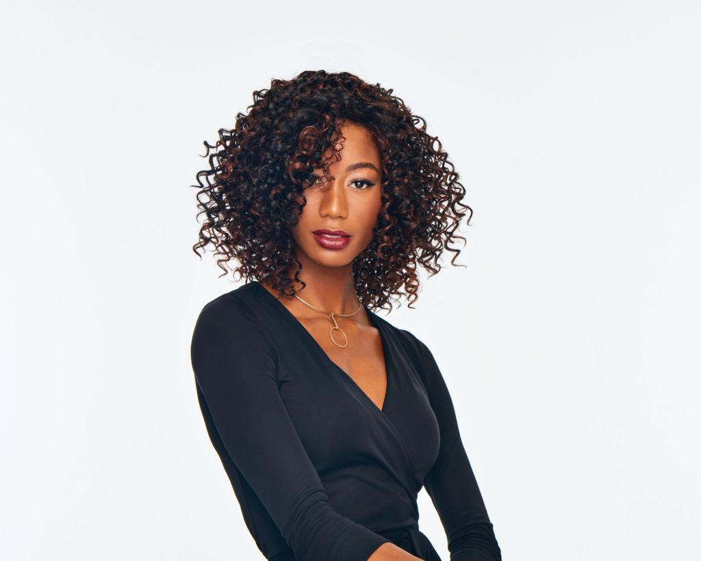 Woman with curly wig and black outfit, looking confident.