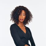 Woman with curly wig and black outfit, looking confident.