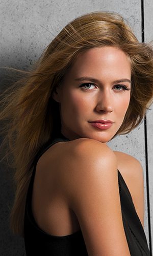 Close-up of a woman with long, wavy blonde wig against a concrete wall.
