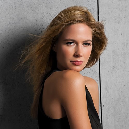 Close-up of a woman with long, wavy blonde wig against a concrete wall.