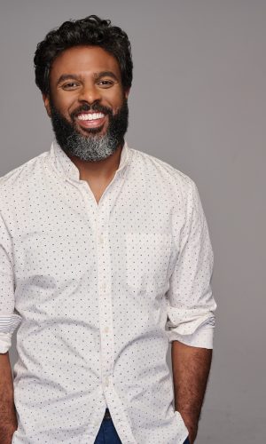 Smiling man with curly hair, beard, and white shirt against gray background.