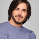 Man with styled dark brown wig, casual blue shirt, confident expression.