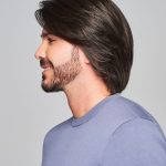 Side profile of a man with a sleek, dark brown wig and beard, smiling confidently.