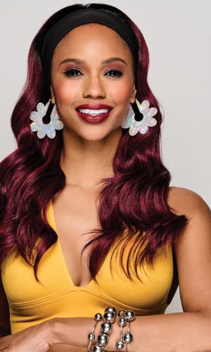Woman with wavy burgundy wig, headband, floral earrings, and yellow top.