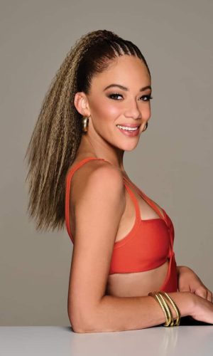Model with crimped pocket ponytail wig, smiling, wearing orange top and gold jewelry.
