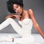 Model wearing a curly black wig with white outfit and gold jewelry.