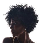 Black curly wig styled on a woman with gold jewelry and earrings.