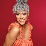 Woman with silver curly wig styled in a high bun and bangs, smiling against a red background.