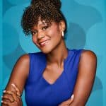 Woman with curly hair styled in a bun and bangs, wearing a blue top and gold jewelry.