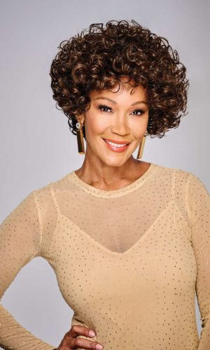 Woman wearing a curly wig with a beige dress and gold earrings.