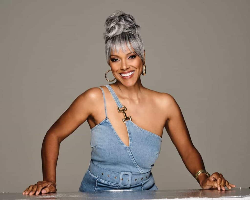 Woman with silver bun and bangs wearing denim top and gold accessories.