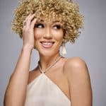 Woman with curly blonde wig, smiling, wearing a beige dress and jewelry.