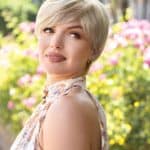 Short blonde wig styled on a woman outdoors with pink flowers in background.