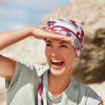 Smiling woman in a colorful headscarf shielding her eyes at the beach.