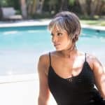Woman with short gray wig by poolside in sunlight.