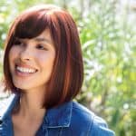 Woman with auburn bob wig outdoors, smiling, natural sunlight, lush green background.