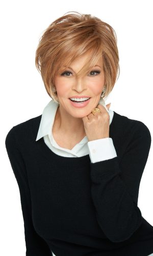 Woman with short auburn wig, smiling, wearing a black sweater and white shirt.