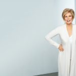 Smiling woman with short blonde wig and white outfit in bright room.