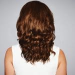 Back view of a woman with wavy auburn hair wearing a white top.
