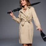 Woman in beige trench coat holding black umbrella and purse, smiling confidently.