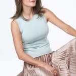 Woman with shoulder-length brown wig wearing a light blue top and beige pleated skirt.