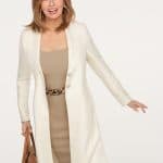 Woman with shoulder-length hair wearing beige dress and white coat, holding a brown handbag.