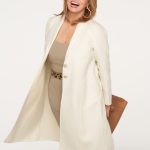 Woman smiling in cream coat and beige dress, holding a brown clutch.