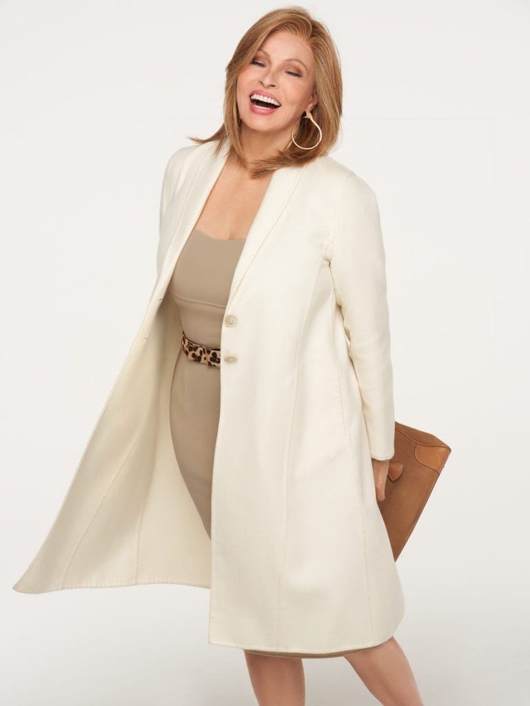 Woman smiling in cream coat and beige dress, holding a brown clutch.
