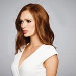 Woman with auburn wavy wig and white top, looking to the side.