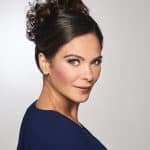 Woman with dark brown curly updo wig and blue top, looking at camera.