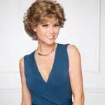 Woman with short, curly wig in a blue dress smiling at camera.